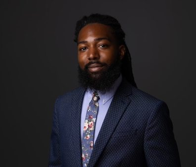 Headshot of a man with a beard in a navy suit, light blue shirt, and floral tie