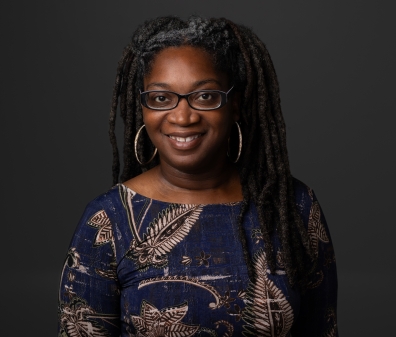 Headshot of a woman with glasses and dark dreadlocks wearing a navy shirt with a beige floral leaf pattern