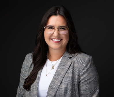 Headshot of a woman in glasses with long brown hair, a white shirt, and a gray plaid blazer