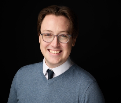 Headshot of a man with glasses wearing a grey sweater, white collared shirt, and dark tie